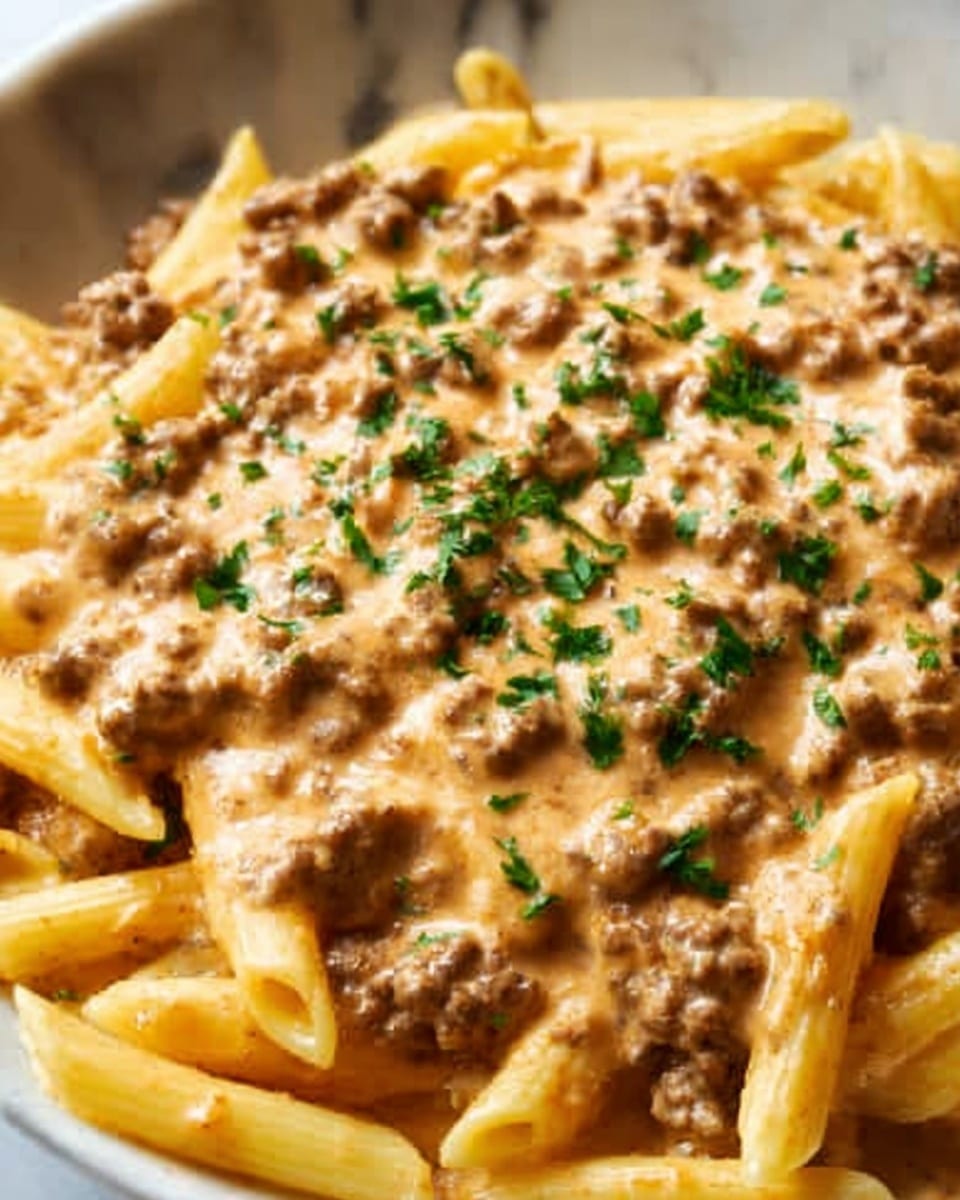 The image shows a close-up of a creamy pasta dish with penne pasta coated in an orange-tan sauce that has a smooth and slightly thick texture. Mixed throughout the pasta are small pieces of ground meat that add a crumbly texture, and the dish is topped with small green parsley leaves scattered evenly, adding a touch of color contrast. The pasta sits on a white plate, and the background is a white marbled surface, giving a clean and bright look. The overall appearance is rich and appetizing, highlighting the sauce's creaminess and the pasta's softness. Photo taken with an iphone --ar 4:5 --v 7
