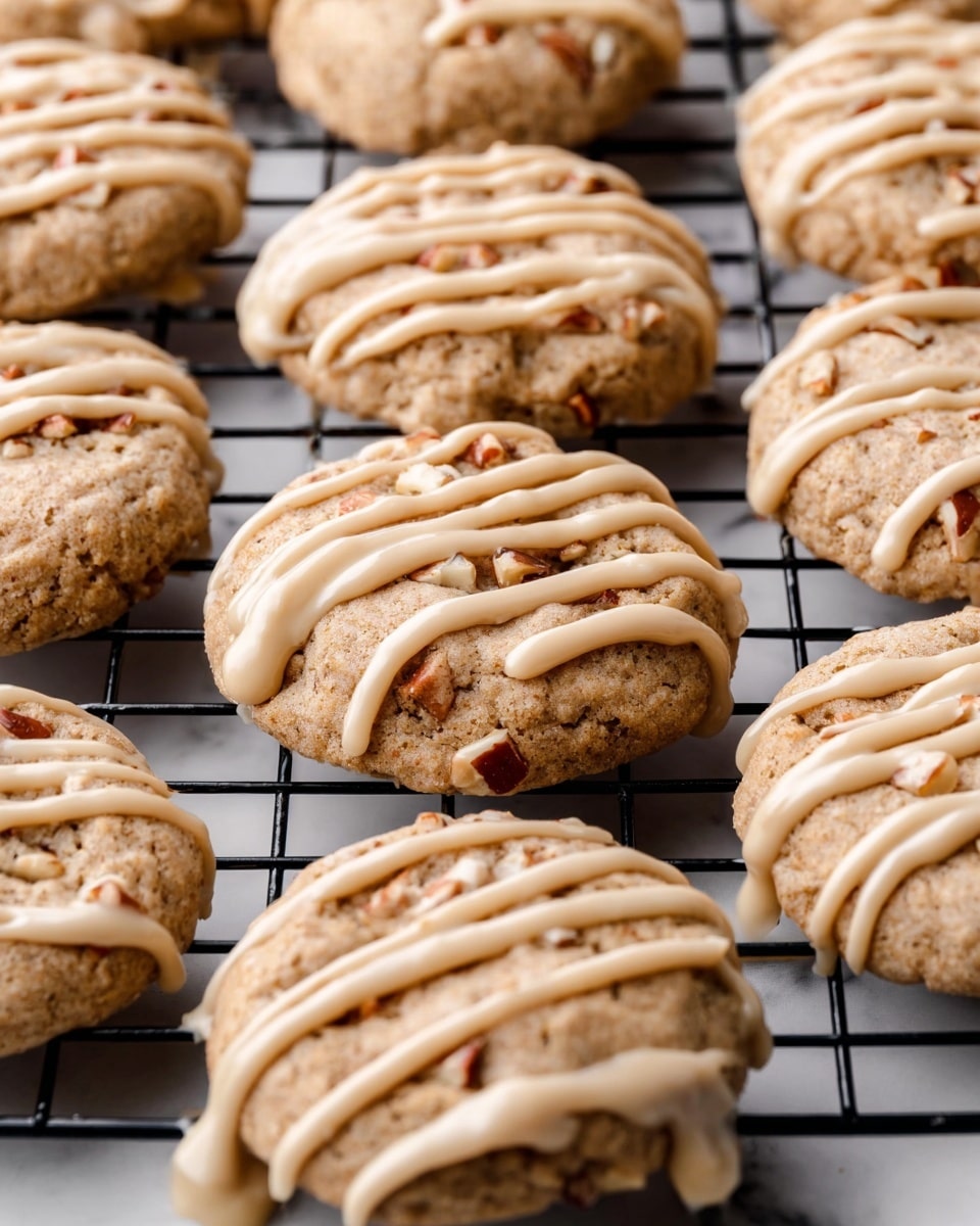The image shows a close-up of multiple round cookies sitting on a black wire cooling rack placed on a white marbled surface. Each cookie has a light brown color with visible small bits of nuts inside, giving a slightly rough texture. On top of every cookie, there is a drizzle of creamy beige icing applied in uneven stripes that lightly spills over the edges, creating small drops on the rack beneath. The cookies appear soft and thick, each about the same size and arranged close together, filling the frame. Photo taken with an iphone --ar 4:5 --v 7
