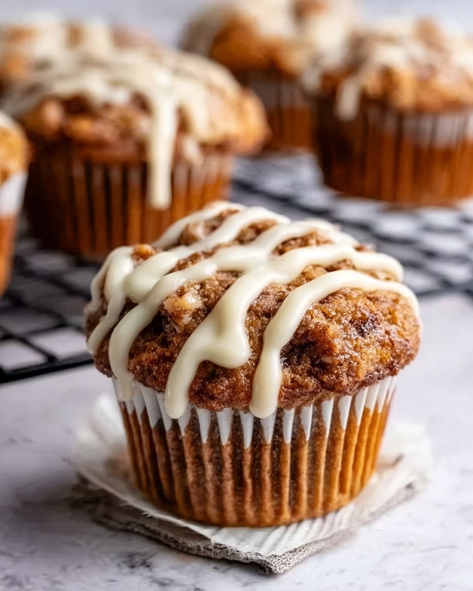 The image shows a close-up of a single muffin on a white paper liner with brown and golden textures, topped with a thick drizzle of cream-colored icing. The muffin’s surface is rough with visible bits of nuts or fruit, giving it a crunchy look. In the background, there are more muffins with similar texture and icing, slightly out of focus on a black wire rack. The whole setup is on a white marbled surface. photo taken with an iphone --ar 4:5 --v 7