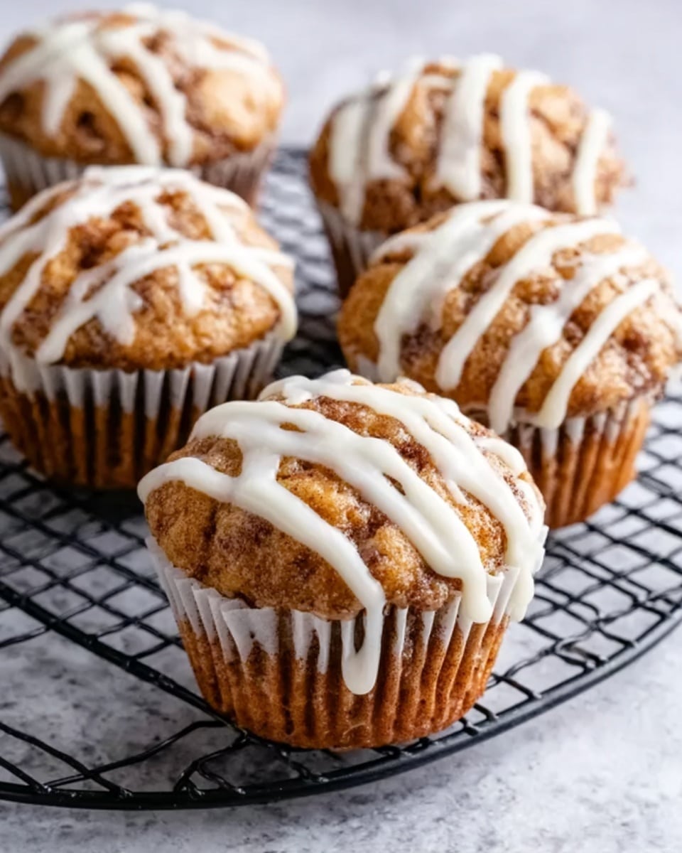 A close-up view of six cinnamon muffins arranged on a black round cooling rack, each muffin topped with a thick layer of white icing drizzled in uneven lines, giving a glossy and creamy texture. The muffins have a golden-brown color with a slightly crumbly surface showing specks of cinnamon and small folds at the edges, wrapped in white paper liners. The background is a white marbled texture that contrasts softly with the warm tones of the muffins. Photo taken with an iphone --ar 4:5 --v 7