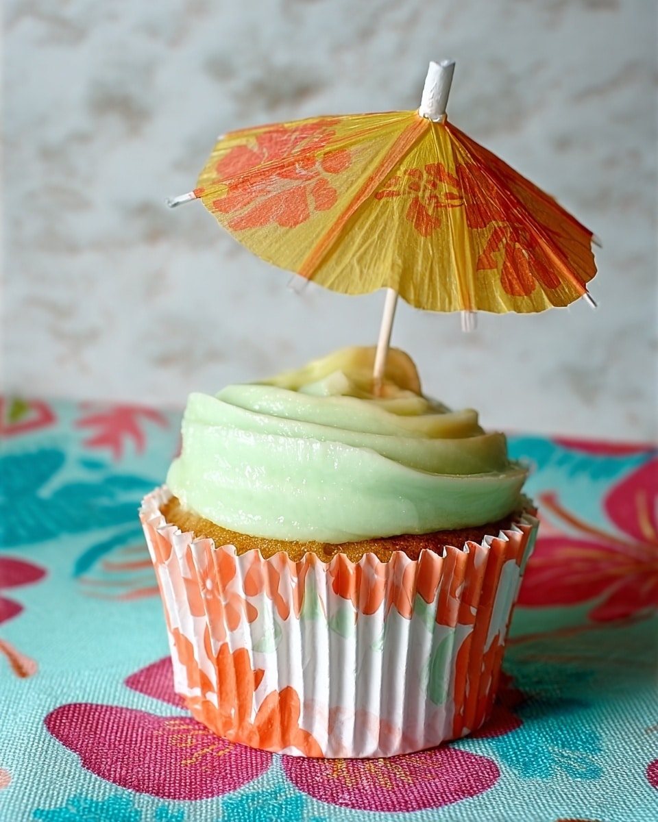 A single cupcake is shown in a white cupcake liner with orange and red flower patterns. The cupcake has one thick layer of pale green frosting on top, smooth and slightly shiny. A small paper umbrella is stuck into the frosting, with an orange and yellow shaded canopy and a white tip, standing upright. The cupcake sits on a white marbled textured surface with a colorful cloth underneath that features pink, blue, and red flower patterns. photo taken with an iphone --ar 4:5 --v 7