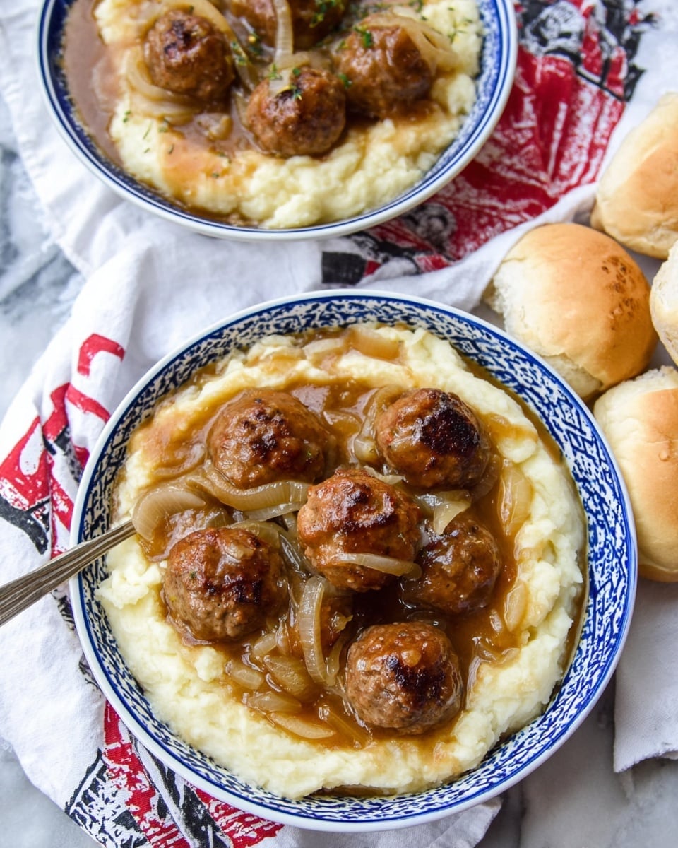 The image shows two white bowls with blue patterns around the edges, each filled with a base layer of creamy mashed potatoes, thick and smooth with a pale yellow color. On top of the mashed potatoes, there are several browned meatballs, round and evenly cooked. The meatballs are covered with a rich brown gravy that has translucent cooked onion slices scattered across it, adding texture and depth. Around the bowls, there are soft, light golden dinner rolls placed on a white marbled surface with a white cloth featuring red and black designs beneath the bowls. The overall look is warm, hearty, and inviting. photo taken with an iphone --ar 4:5 --v 7