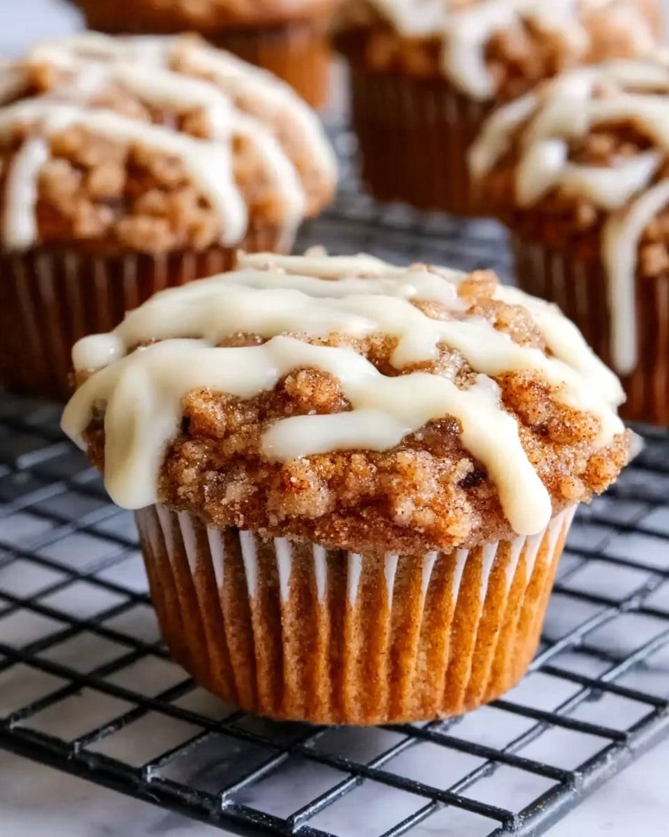 A close-up view of a cinnamon crumb muffin with a crumbly brown top layer and white icing drizzled over it, placed in a white paper muffin liner on a dark wire cooling rack. Behind it, several more muffins are slightly blurred. The background features a soft white marbled texture. The lighting highlights the texture of the crumb topping and smooth icing, making the muffin look fresh and inviting. Photo taken with an iphone --ar 4:5 --v 7