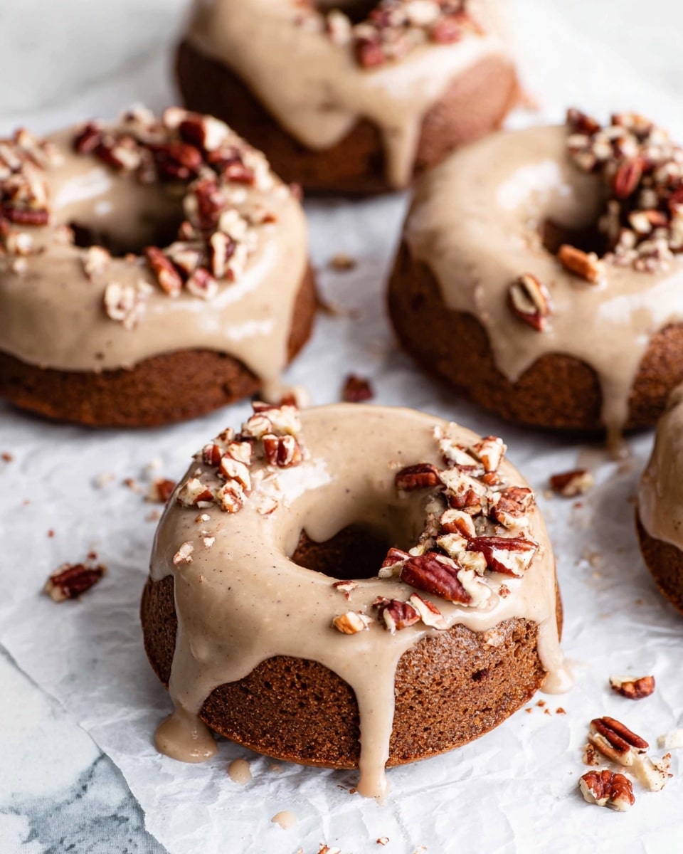 The image shows several round donuts with a hole in the center, each topped with a smooth, light brown glaze that drips slightly over the edges. The donuts have a rich, dark brown texture underneath the glaze, indicating a dense and moist cake. On top of the glaze, some donuts are sprinkled with small, chopped pecan pieces scattered unevenly, adding texture and contrast with their rich reddish-brown and cream colors. The donuts are placed on a crinkled white paper over a white marbled surface, with a few pecan pieces scattered casually around them. photo taken with an iphone --ar 4:5 --v 7