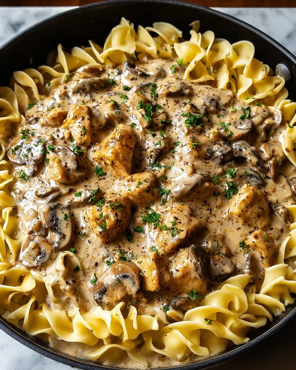 A close-up view of a black skillet filled with creamy chicken stroganoff over egg noodles. The dish has a bottom layer of wide, wavy, pale yellow egg noodles, topped with a thick layer of golden-brown cooked chicken chunks mixed with sliced brown mushrooms. The creamy sauce is light brown with visible black pepper and herb specks, coating the chicken and noodles evenly. Small bits of chopped green parsley are sprinkled on top, adding a fresh touch. The skillet sits on a white marbled surface. Photo taken with an iphone --ar 4:5 --v 7