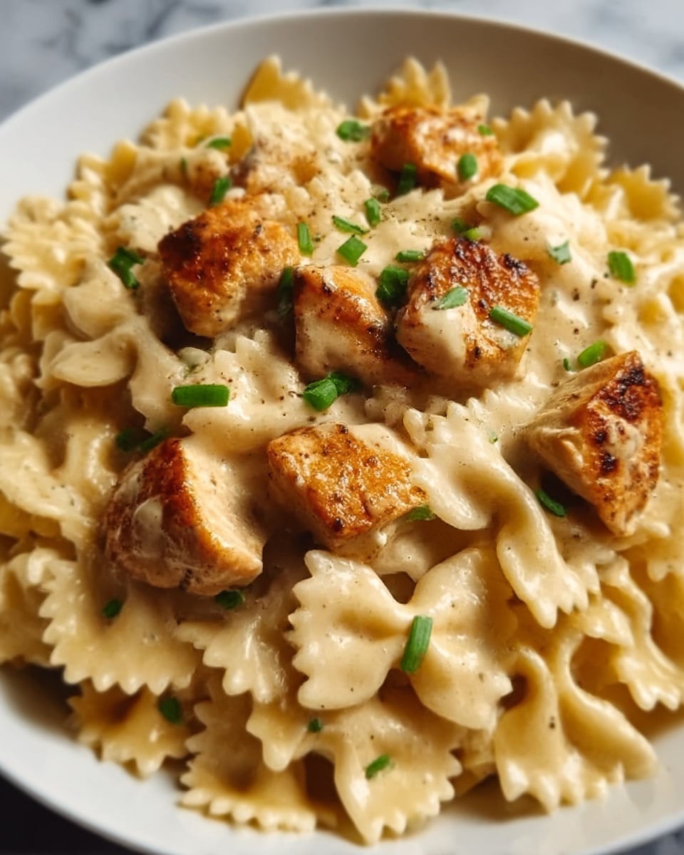 The image shows a close-up of a white plate filled with bowtie pasta coated in a creamy, light beige sauce. On top, several browned chicken pieces rest scattered evenly, with a slightly crispy texture. Small green herbs are sprinkled over the dish, adding a touch of fresh color. The creamy sauce appears smooth and thick, covering the pasta without dripping. The background is a white marbled surface. Photo taken with an iphone --ar 4:5 --v 7