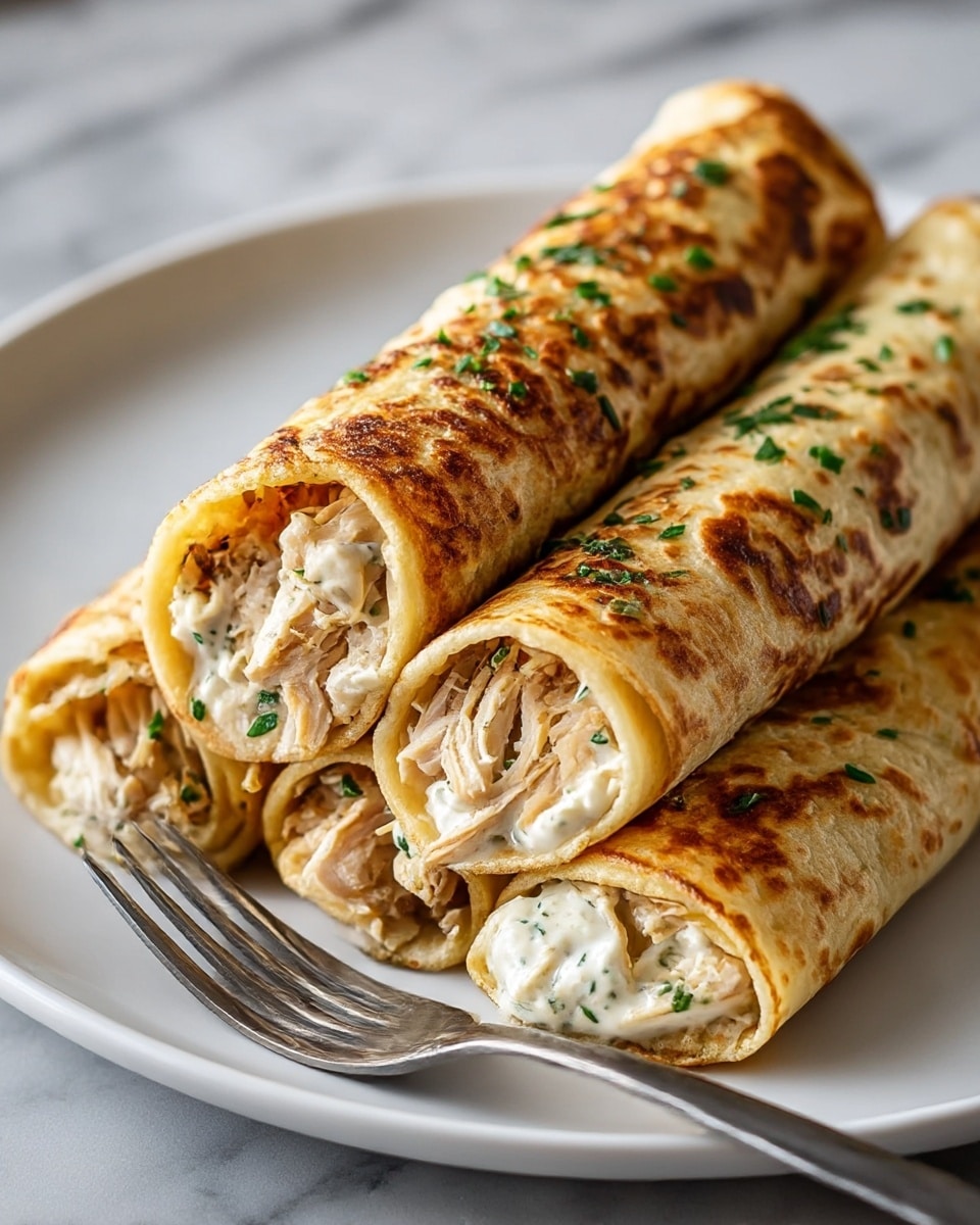 This image shows a white plate holding four rolled crepes stacked close together. Each crepe is golden brown with a slightly crispy outer layer, speckled with darker toasted spots, and sprinkled with chopped green herbs on top. Inside, the crepes are filled with two layers: the first layer is shredded, light brown chicken, and the second layer is a creamy white sauce with small green herb bits mixed in. A silver fork rests on the edge of the plate. The plate sits on a white marbled surface. photo taken with an iphone --ar 4:5 --v 7