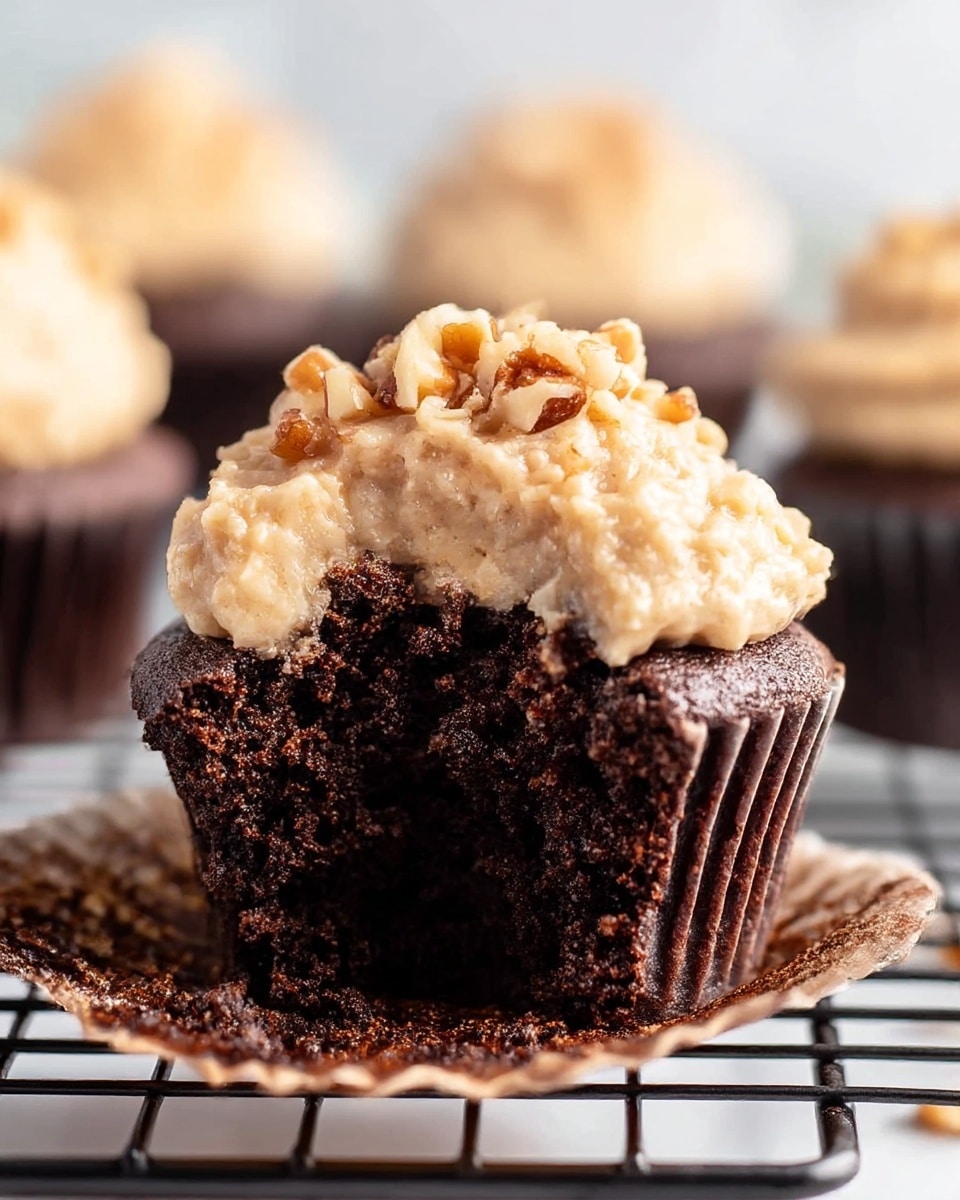A close-up view of a chocolate cupcake with one bite taken out, showing the dark, moist, and crumbly texture of the cake. On top is a thick layer of light brown frosting with a rough texture, sprinkled with small chunks of nuts. Behind the main cupcake, there are blurred similar cupcakes which also have the nutty frosting. The cupcakes rest on a cooling rack placed on a white marbled surface. photo taken with an iphone --ar 4:5 --v 7