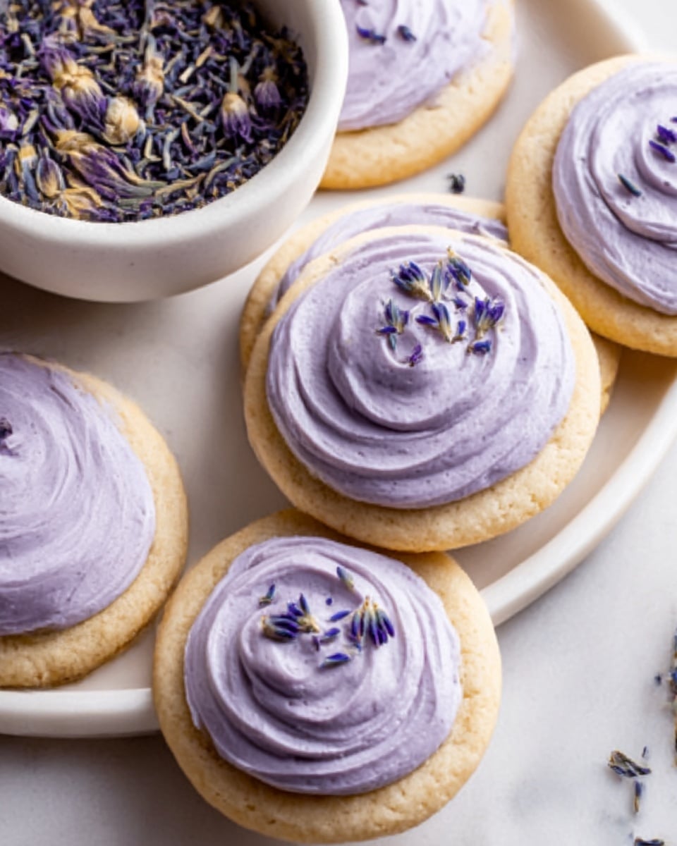 The image shows several round cookies with soft, light tan bases topped with a thick layer of light purple frosting, swirled smoothly in circular motions. Each cookie has a small amount of dried lavender sprinkled on top of the frosting. The cookies rest on a white oval plate placed on a white marbled surface. Nearby, there is a small white bowl filled with dried lavender flowers. A white flower is partially visible near the bottom edge of the image. Photo taken with an iphone --ar 4:5 --v 7